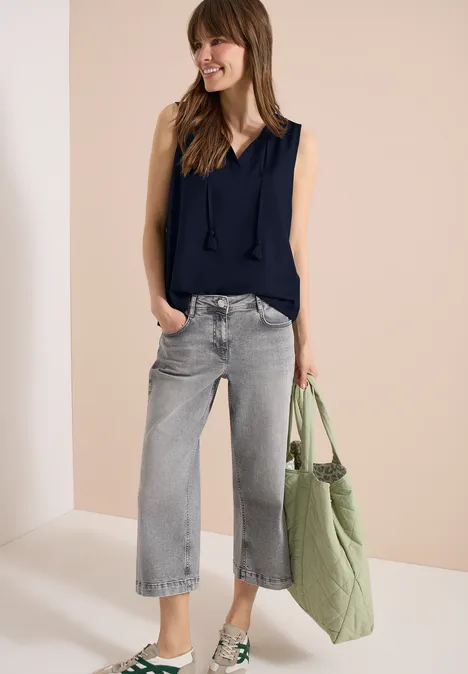 Frau in ärmelloser dunkelblauer Bluse, weiten grauen Jeans und grüner Stofftasche vor beigem Hintergrund.