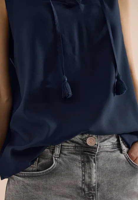 Person trägt dunkelblaue Bluse mit Quasten und hellgraue Jeans, Hände in den Taschen.