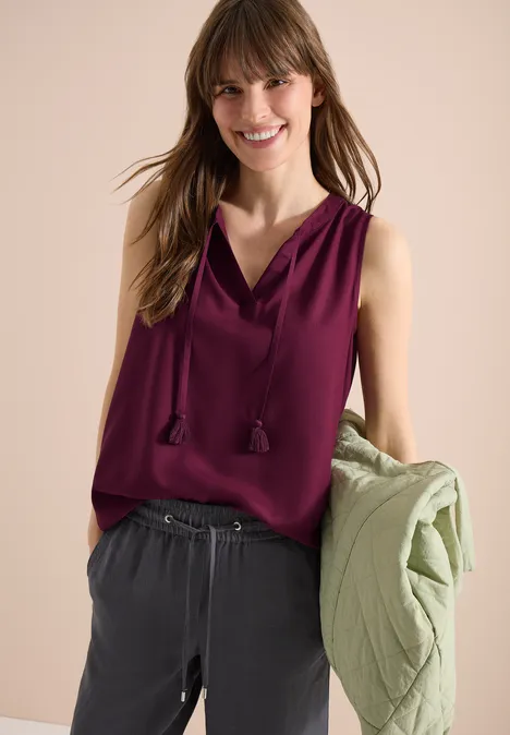 Une femme porte un haut sans manches bordeaux, un pantalon gris et tient une veste verte