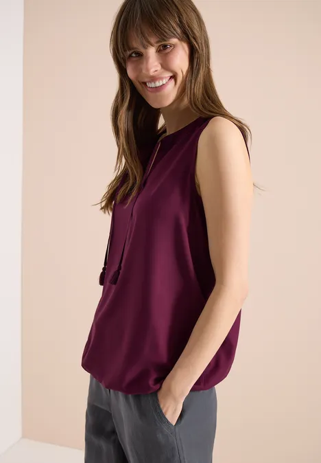 Femme en top sans manches violet foncé et pantalon gris, main dans la poche, fond clair