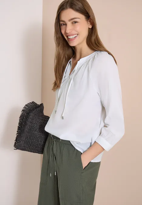 Frau trägt weiße Bluse, olivgrüne Hose und hält schwarze geflochtene Clutch, steht vor beigem Hintergrund.