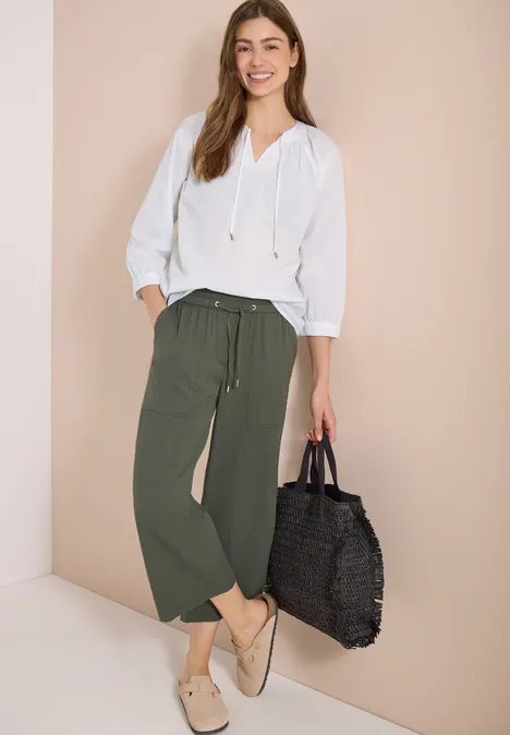 Frau trägt weiße Bluse, lockere grüne Hose, beige Slipper und schwarze Basttasche.