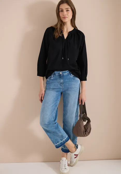 Person in schwarzer Bluse, blauen Jeans und weißen Sneakern steht mit grauer Tasche vor beige Wand.