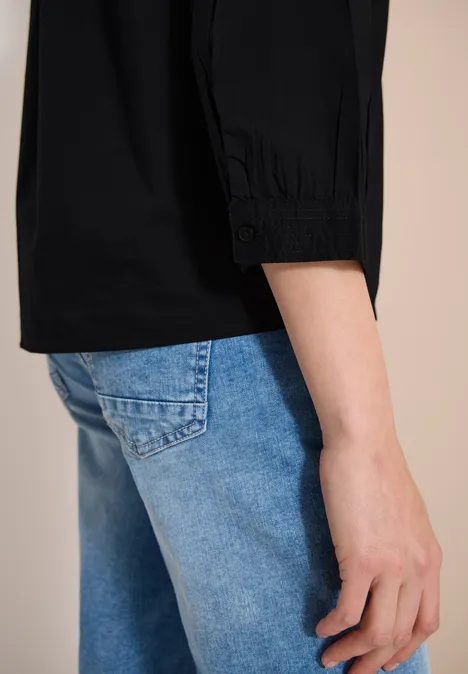 Eine Person trägt eine schwarze Bluse mit 3/4-Ärmeln und hellblaue Jeans, seitliche Nahaufnahme.