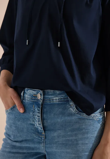Frau trägt dunkelblaue Bluse und Jeans; eine Hand steckt lässig in der Hosentasche.
