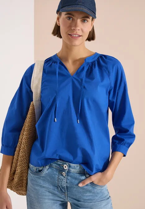 Persoon draagt blauwe blouse met koorden, spijkerbroek en tas voor een neutrale achtergrond.