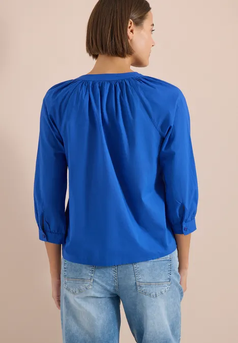 Vrouw van achteren in een blauw 3/4-mouwen top met plooien en lichte jeans.