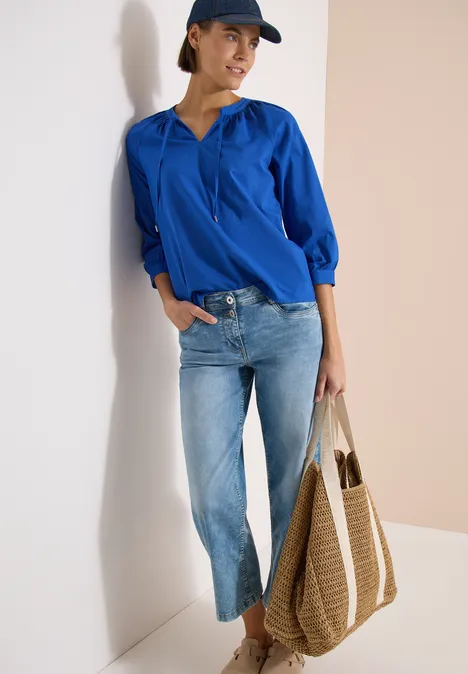 Vrouw leunt tegen muur, draagt blauw overhemd, lichte jeans, pet en grote gevlochten shopper.