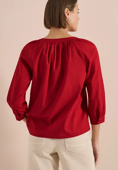 Frau von hinten in roter, locker sitzender Bluse und hellen Hosen vor hellem Hintergrund.