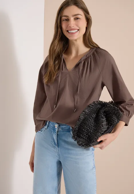 Frau mit brauner Bluse und Jeans hält schwarze Clutch vor hellem Hintergrund.