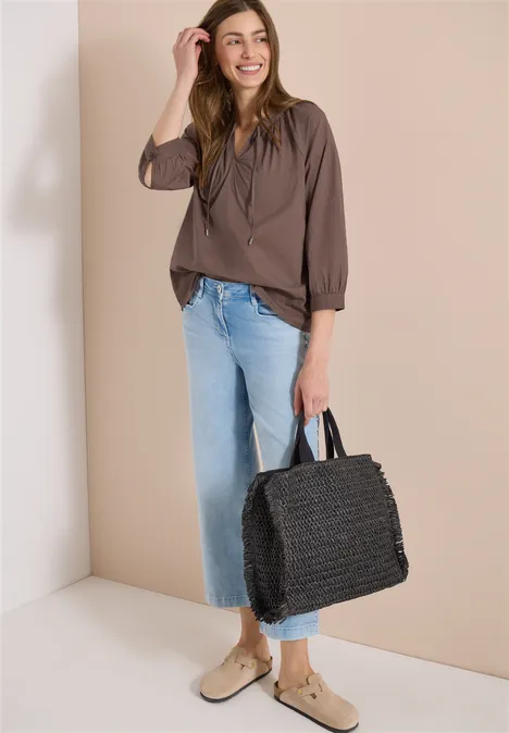 Frau trägt braune Bluse, helle Jeans, beigen Schuhen und hält eine schwarze Korbtasche.