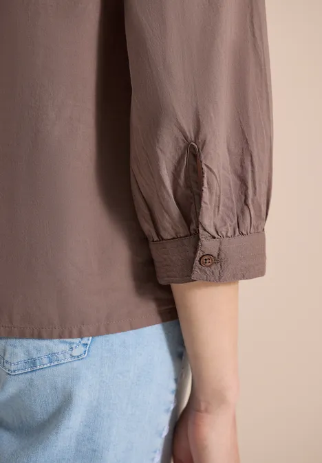 Ein Arm in taupefarbener Bluse mit hochgekrempeltem Ärmel und hellblauer Jeans vor hellem Hintergrund.