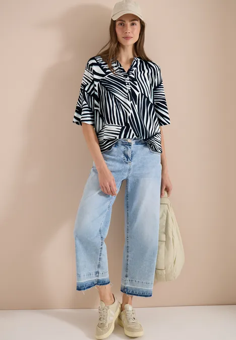Frau in gemusterter Bluse, weiten Jeans und Sneakers, hält eine Tasche.
