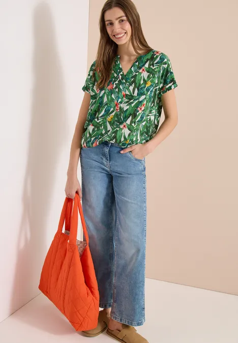 Vrouw in groene blouse met bladmotief, wijd jeans en oranje tas tegen neutrale achtergrond.