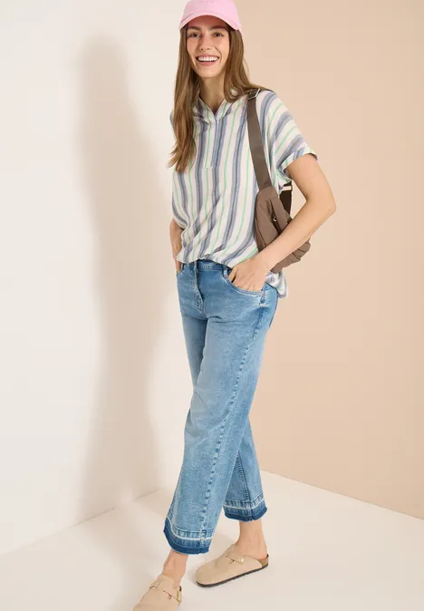 Kurzarm Bluse mit Split Neck und Streifen vanilla white Frau trägt gestreiftes Hemd, weite blaue Jeans und bequeme Hausschuhe, hält eine Tasche, steht vor hellem Hintergrund.