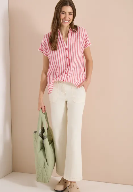 Frau trägt rot-weiß gestreiftes Hemd, weiße Hose und hält eine grüne Tasche vor beiger Wand.