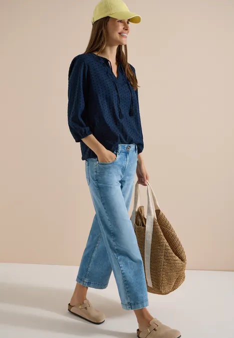 Vrouw in casual outfit: donkerblouse, wijde spijkerbroek, pet, gevlochten tas en sandalen.