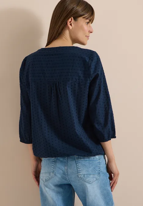 Rugaanzicht van een vrouw in een donkerblauwe blouse met subtiel stippenpatroon en een lichte spijkerbroek.