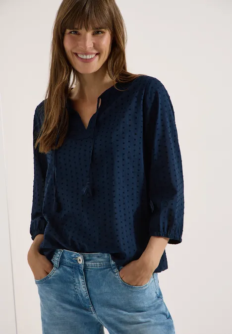 Vrouw in donkerblauw, subtiel dessinblouse met V-hals en spijkerbroek, handen ontspannen in de zakken.