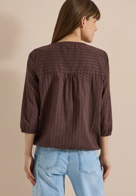 Achteraanzicht van een vrouw in een bruin shirt met stippen, driekwartsmouwen en een lichtblauwe spijkerbroek.
