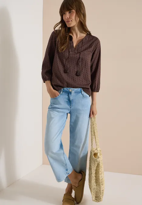 Vrouw in bruin bloesje, lichte wijde jeans en loafers, met een grote gevlochten strottas.