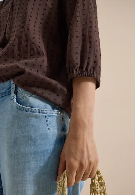 Een persoon in lichtblauwe jeans en bruine blouse houdt een beige, met kralen versierd handtasje vast.