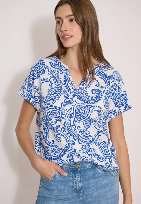 Blouse met split in de hals en print vanilla white Frau trägt weiß-blau gemustertes V-Neck-T-Shirt und Jeans, Hand in Hosentasche, vor neutralem Hintergrund.