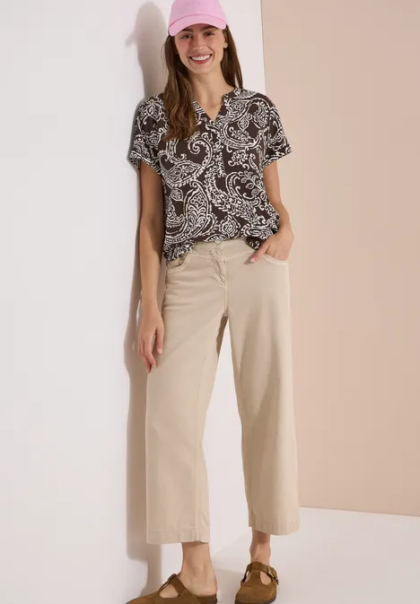 Blouse met split in de hals en print brown cacao Frau lehnt an zweifarbigem Hintergrund, trägt gemusterte Bluse, weite Beige-Hosen und Slingback-Pumps.