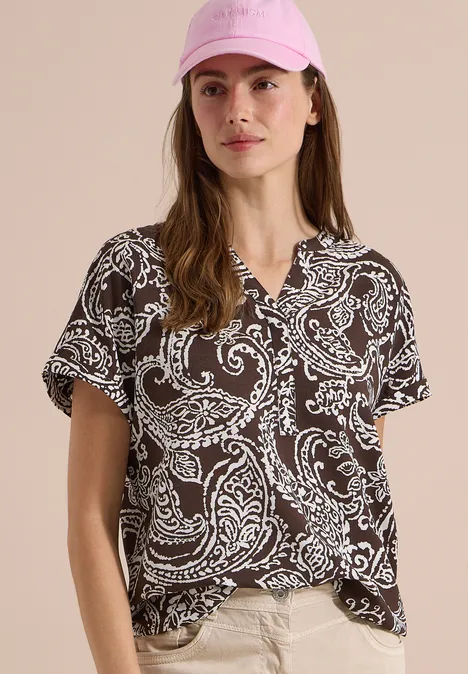 Blouse met split in de hals en print brown cacao Frau in braunem T-Shirt mit weißem Paisley-Muster, kurzen Ärmeln und V-Ausschnitt vor neutralem Hintergrund.