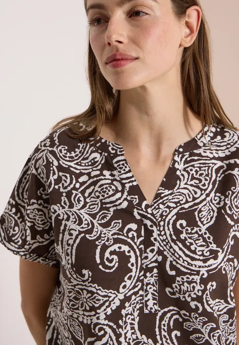 Blouse met split in de hals en print brown cacao Frau trägt eine kurzärmelige, dunkelbraune Bluse mit weißem, paisleyartigem Muster.