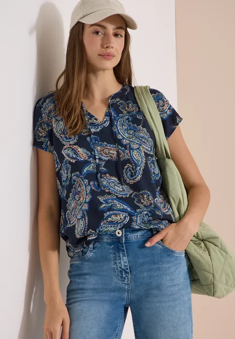 Blouse met split in de hals en een mix van texturen universal blue Frau mit blau-grüner Paisley-Bluse, Jeans und grüner gesteppter Tasche lehnt an der Wand.
