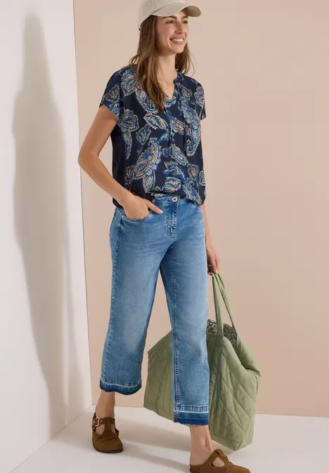 Blouse met split in de hals en een mix van texturen universal blue Frau im beigen Cap, trägt gemusterte Kurzarmbluse, weite Cropped-Jeans und grüne Stepp-Shopper-Tasche.