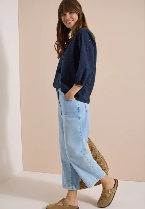 Eine Frau trägt eine dunkelblaue Jacke, helle Jeans, Basttasche und beige Loafers.