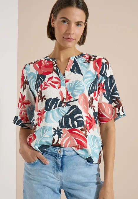 Kurzarm Bluse mit Split Neck light alabaster white Frau trägt bunte Bluse mit tropischem Blattmuster und helle Jeans.