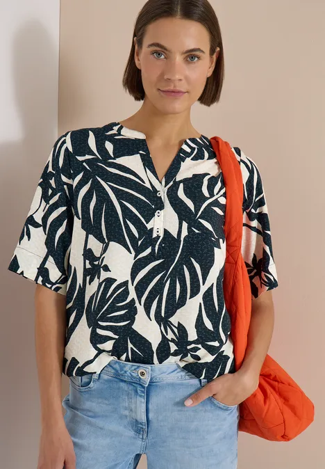 Blouse met korte mouwen en gespleten hals palm breeze green Frau mit schwarz-weißem Blusen-Shirt mit Blattmuster, Jeans und orangefarbener Tasche vor beigem Hintergrund.
