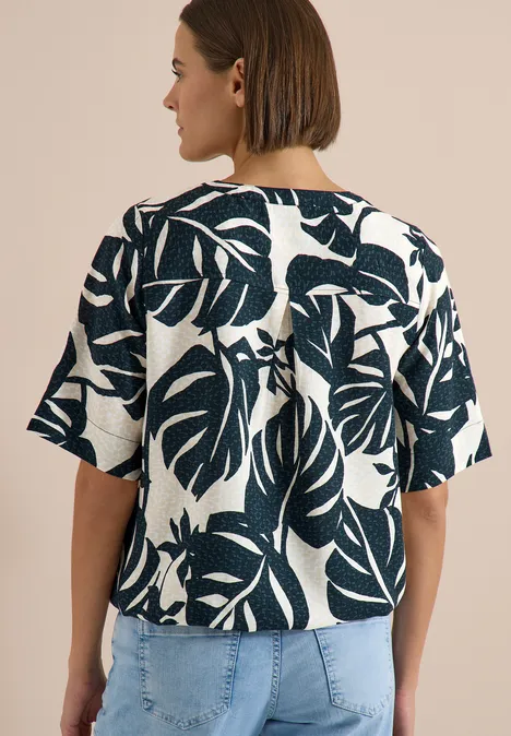 Blouse met korte mouwen en gespleten hals palm breeze green Frau von hinten in Jeans und weißem, kurzärmligem Oberteil mit großem dunkelgrünem Blattmuster.