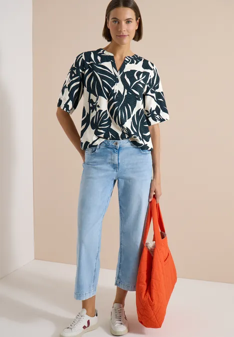 Blouse met korte mouwen en gespleten hals palm breeze green Frau trägt schwarz-weißes Blattmuster-Hemd, hellblaue Jeans, weiße Sneaker und hält eine orangefarbene Tasche.