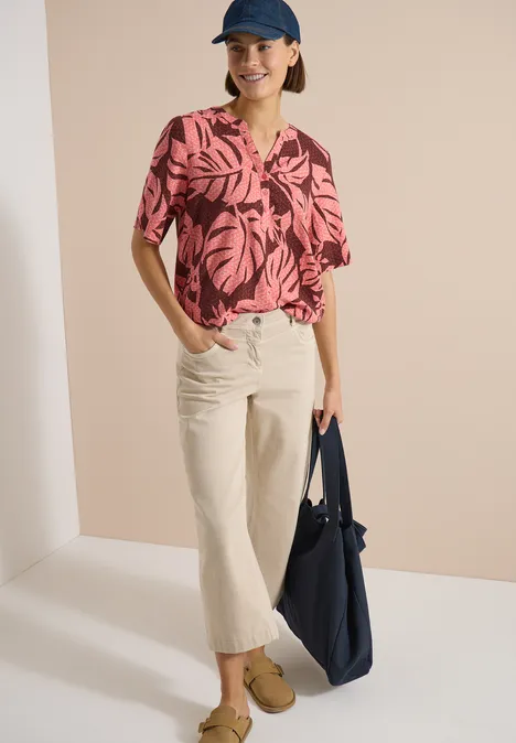 Blouse met korte mouwen en gespleten hals null Model trägt rosa-schwarze Bluse mit Blattmuster, helle Culotte, beige Schuhe, blaue Tasche und Mütze.