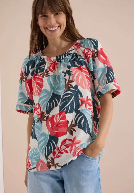 Frau trägt eine weite Bluse mit buntem Blumen- und Blattmuster zu hellblauen Jeans.