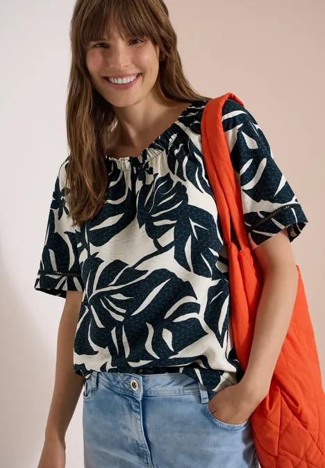 Frau trägt Jeans, weißes kurzärmeliges Shirt mit dunkelblauem Blattprint und eine rote Tasche über der Schulter.