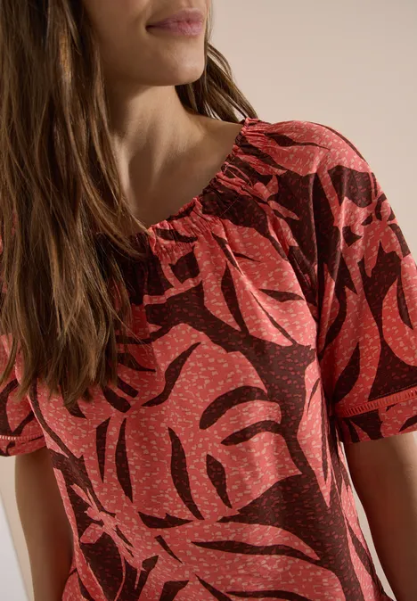 Carmen-Bluse mit Kurzarm und Blätterprint coral Frau mit braunen Haaren trägt rotes, braun gemustertes kurzärmeliges Oberteil, Nahaufnahme des Oberkörpers.
