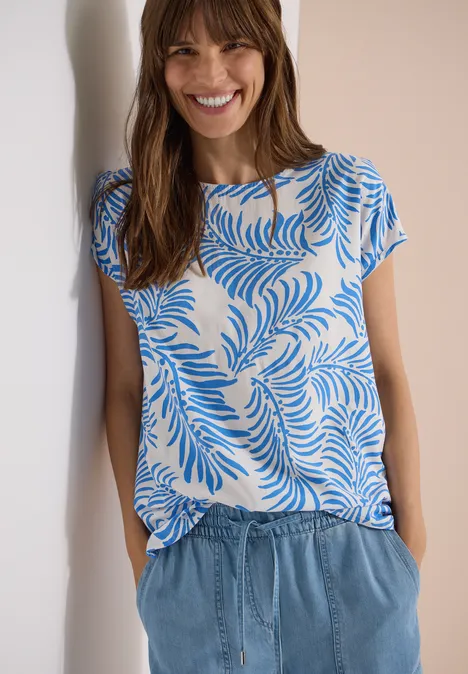 Frau im weißen T-Shirt mit blauem Blattmuster und blauen Jeans, Hände in den Taschen.