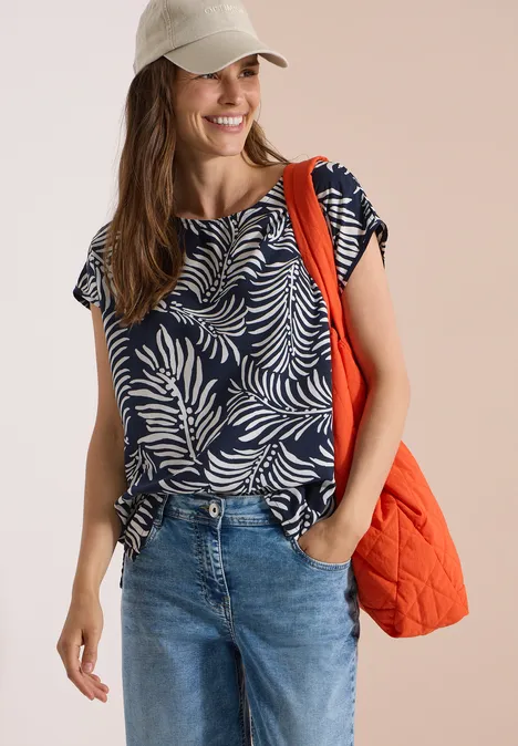 Frau in Jeans, navyblauem Blattmuster-Shirt und Cap trägt eine orangefarbene gesteppte Tasche.