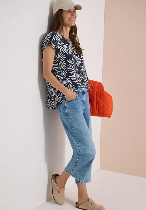 Frau lehnt an Wand, trägt gemustertes Top, weite Jeans, bequeme Schuhe und hält eine orange Tasche.