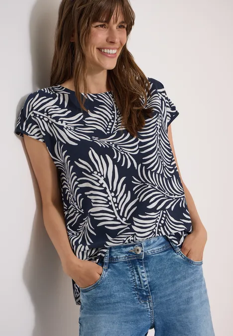 Eine Person trägt ein dunkelblaues Shirt mit weißem Blattmuster und Jeans und hat die Hände in den Taschen.