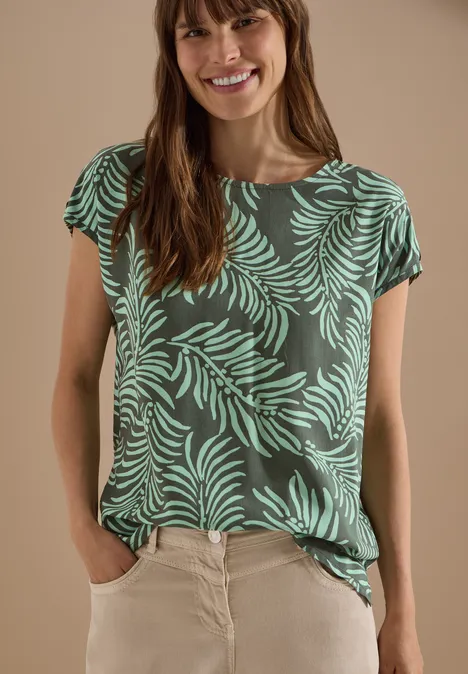 Eine Frau mit verschwommenem Gesicht trägt ein grünes T-Shirt mit Blattmuster und helle Hosen.