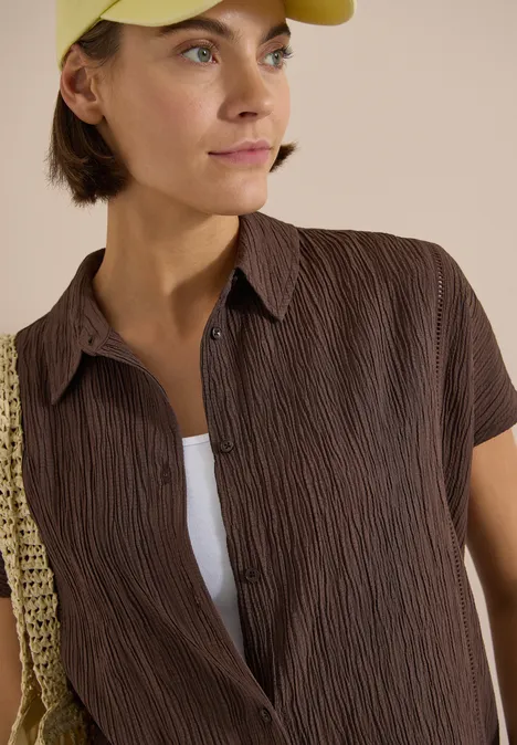 Dropped Shoulder Hemdbluse in Unifarbe brown cacao Frau trägt braune, kurzärmlige, strukturierte Bluse über weißem Top und hält eine gehäkelte Strohtasche.