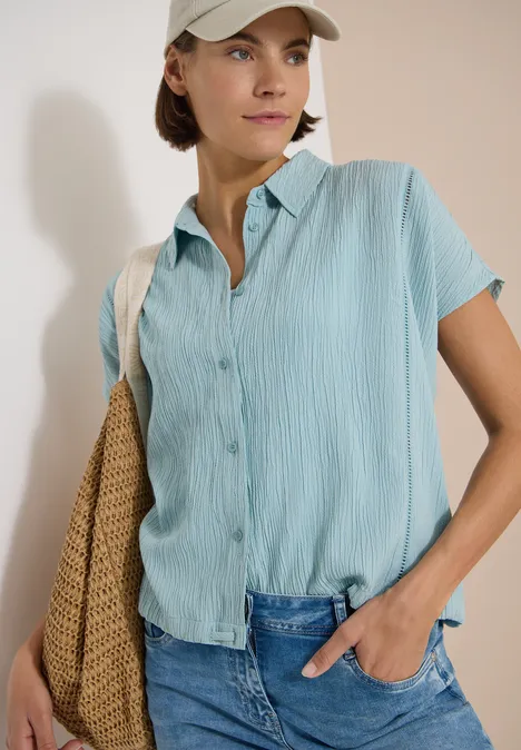 Dropped Shoulder Hemdbluse in Unifarbe crystal mint Frau mit hellblauer Kurzarmbluse, blauen Jeans und geflochtener Strohtasche lehnt an weißer Wand.