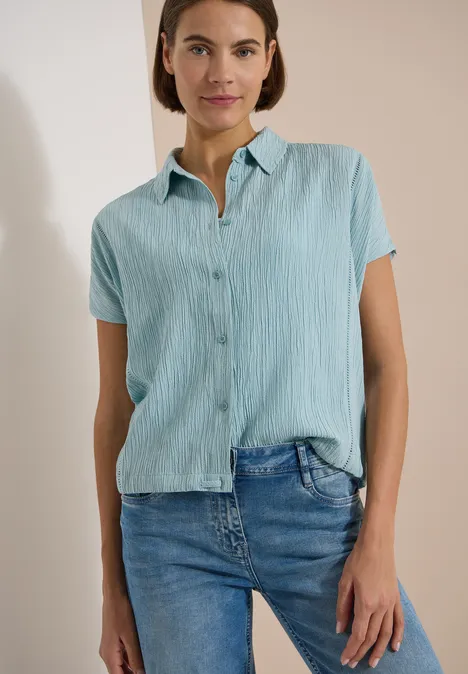 Dropped Shoulder Hemdbluse in Unifarbe crystal mint Eine Frau trägt eine hellblaue, kurzärmelige Bluse und Jeans vor einem ruhigen Hintergrund.