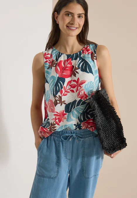 Blouse met ronde hals en bladerenprint light alabaster white Vrouw draagt mouwloos shirt met rood bloemmotief, blauwe broek en houdt een zwarte tas vast.